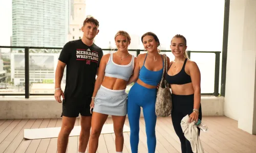 Four people stand together on a wooden terrace with a cityscape in the background. The group consists of one man and three women, all dressed in athletic wear. The first woman wears a light blue sports bra and matching skirt, the second woman is in a bright blue sports bra and leggings, and the third woman wears a black sports bra and leggings, holding a water bottle and a towel. The setting appears to be outdoors or on a rooftop, with tall buildings visible behind them. The atmosphere is casual and sporty