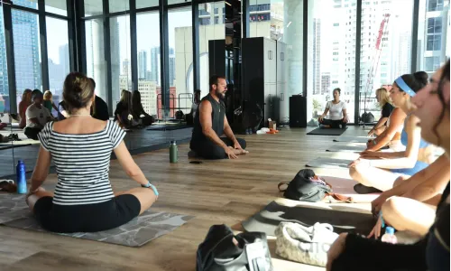 A group of people practicing yoga in a bright studio with floor-to-ceiling windows overlooking modern Miami skyscrapers. The instructor sits in the center of the room on a yoga mat in a cross-legged position, while participants mirror the pose on their own mats arranged in a circle. The space is filled with natural light, and there are gym bags and water bottles on the wooden floor. The atmosphere is calm and focused, emphasizing health and harmony